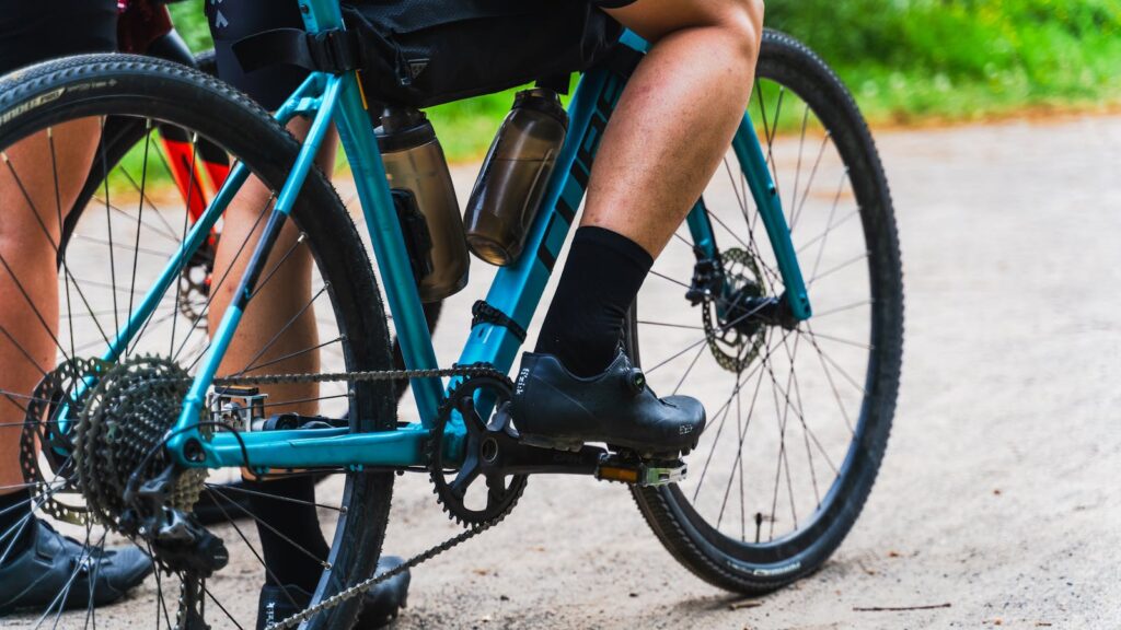 Close-up of a cyclist's legs and bicycle on a gravel path. Sports gear and water bottles visible.
